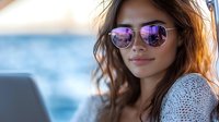 Young Asian woman in sunglasses with purple reflection smiling at sunset, wearing white knit sweater against blurred ocean background, natural warm lighting.