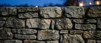 Ancient stone wall with weathered rocks stacked in traditional masonry pattern during twilight hour with warm lights glowing softly in blurred background.