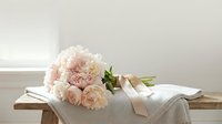 Soft blush peony bouquet tied with a satin ribbon resting on a linen blanket on a rustic wooden bench in a bright airy room.