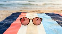 Stylish round sunglasses with rose gold mirrored lenses resting on colorful striped beach towel with ocean waves and sandy shore in soft focus background.