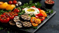 Grilled vegetables platter with eggplant, bell peppers, tomatoes, and creamy dip, garnished with herbs on dark slate board.