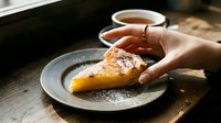 Slice of custard tart dusted with powdered sugar on a rustic plate, hand reaching for it, warm cup of tea in background.