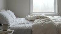 Unmade bed with white linens, pillows, and rumpled duvet near bright window with natural daylight streaming in, suitable for bedroom, lifestyle, and home decor designs.