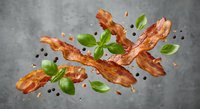 Crispy bacon strips with fresh basil leaves and black peppercorns flying in motion against gray concrete background for culinary design projects.