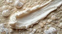 Creamy white cosmetic product smeared on sandy beige texture with scattered droplets, showcasing skincare or makeup application for beauty and wellness marketing.