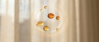 Floating amber glass spheres and droplets suspended in soft natural light with warm beige curtains in background, suitable for cosmetics, skincare, and wellness product marketing.