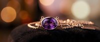 Elegant silver pendant necklace with oval purple amethyst gemstone on delicate chain, displayed on dark stone with warm bokeh background for jewelry retail and luxury accessories.