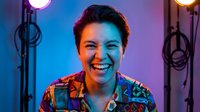 Young Asian woman laughing joyfully in colorful neon studio lighting setup with professional photography equipment for creative portrait sessions.