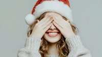 Young Caucasian woman in Santa hat covering eyes with hands, smiling broadly, wearing cozy gray sweater against neutral background.