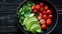 Fresh sliced avocado, cherry tomatoes and baby spinach leaves arranged on black plate against dark wooden background, healthy food composition.