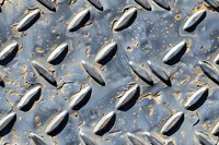 Weathered metal diamond plate with rust and wear pattern showing industrial texture and grip surface detail in gray and orange tones.