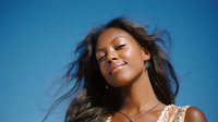 Young African American woman with flowing hair smiling confidently against vibrant blue sky, wearing delicate lace dress and silver jewelry, radiating joy and natural beauty.
