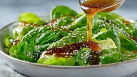 Fresh green salad with sesame seeds being drizzled with golden dressing in white bowl. Healthy vegetarian meal preparation for restaurant menu.