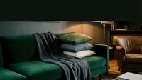 Emerald green velvet sofa with stacked decorative pillows and draped charcoal throw blanket in warm ambient lighting, suitable for interior design and home decor projects.