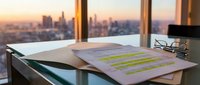 Business documents with highlighted text on modern office desk overlooking city skyline at sunset for corporate analysis and planning.