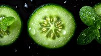 Fresh cucumber slices with water drops and basil leaves on black background. Macro shot of raw organic vegetable showing detailed texture and seeds.
