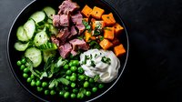 Fresh bowl with rare beef slices, roasted sweet potato cubes, green peas, cucumber, herbs and creamy sauce served on black plate, healthy protein meal.