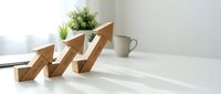 Wooden arrow blocks showing upward growth trend on white desk with green plants and coffee mug in bright office workspace for business success concepts.