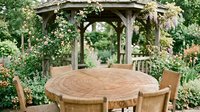 Wooden round dining table with chairs in garden gazebo surrounded by lush green plants and flowering vines, outdoor entertaining space.