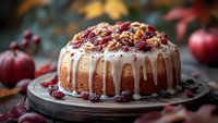 Homemade bundt cake topped with white glaze dripping down sides, garnished with candied cranberries and nuts, served on wooden board with autumn decor.