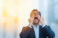 Asian businessman in glasses and suit expressing extreme frustration and stress, screaming with clenched fists against blurred urban background.