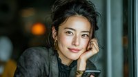 Young Asian businesswoman smiling confidently while looking out office window, hand on chin in thoughtful pose during daytime work break.