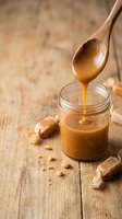 Thick caramel sauce dripping from a wooden spoon into a glass jar, surrounded by wrapped toffee candies on a rustic wooden surface.