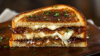 Grilled sandwich with melted cheese and caramelized onions on toasted bread, garnished with fresh herbs, served on wooden cutting board. Close up food photography.