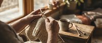 Elderly woman hands knitting warm wool sweater by sunny window with coffee cup and books creating cozy homemade craft atmosphere for relaxation and hobby.