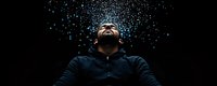 Young man in black jacket looking up at night sky with glowing blue particles falling like stars, expressing wonder and spiritual connection. Panoramic composition.