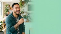 Young adult male with dark hair and facial hair, Hispanic ethnicity, wearing a teal button-up shirt, smiling confidently in a bright, plant-filled interior space with mint green walls.