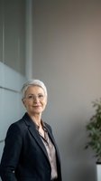 Senior woman with short white hair and glasses wearing dark blazer and light shirt stands in modern office environment with neutral background and green plant.