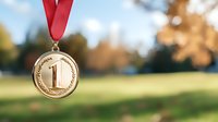 Gold first place medal with red ribbon hanging against blurred natural background with autumn trees and sunlight, symbolizing achievement and victory in sports competition.