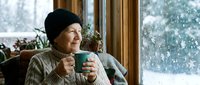 Senior Caucasian woman in cozy sweater and knit hat enjoying hot beverage by window during winter snowfall, creating warm atmosphere for seasonal content.