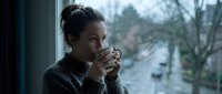 Woman with dark hair in casual sweater drinking hot beverage by rainy window, contemplative mood, suitable for lifestyle and wellness content.