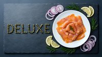 Sliced smoked salmon on white plate with capers spelling deluxe, red onion rings, lemon slices, and dill on dark slate board.
