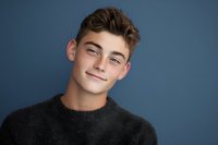 Young handsome Caucasian man with curly brown hair smiling confidently in dark sweater against blue studio background for lifestyle photography.