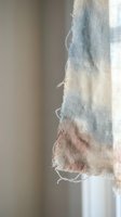 Frayed edge of pastel tie-dye fabric with soft blue and pink colors hanging in natural window light creating artistic textile detail for craft projects.