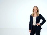 Professional woman with blonde hair wearing dark navy blazer and cream blouse, standing against white background with hand on hip, suitable for corporate portraits and business headshots.