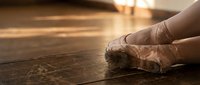 Ballet dancer feet in pointe shoes on wooden floor with warm golden sunlight streaming through windows creating dramatic lighting atmosphere.