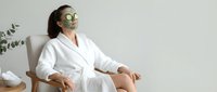 Young woman in white robe relaxing with green clay face mask and cucumber slices, enjoying spa treatment at home wellness routine.