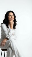 Confident Hispanic businesswoman in white suit sitting on stool with hand on chin, smiling warmly against clean white background for corporate branding.