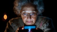 Elderly Caucasian woman with glasses using smartphone at night, blue screen glow illuminating face in dark room for technology and senior lifestyle concepts.