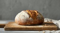 Freshly baked artisan sourdough bread loaf with golden crust on wooden cutting board against concrete wall background for bakery and food photography.