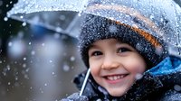 Young Asian child wearing winter hat smiling joyfully while holding clear umbrella during snowfall, capturing pure happiness and wonder of snowy weather outdoors.