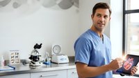 Confident male healthcare professional in blue scrubs holding tablet in modern medical office with microscope and equipment for patient consultation.