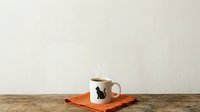Ceramic mug with black cat silhouette design and wooden lid, placed on orange fabric napkin against neutral background. Suitable for product photography, home decor, and kitchenware marketing.
