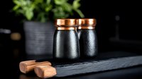 Black ceramic salt and pepper shakers with copper lids on dark slate board against black background. Water drops create elegant texture on matte surface.