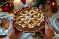 Hands holding homemade apple pie with lattice crust on festive Christmas dinner table with candles and holiday decorations in warm cozy atmosphere.