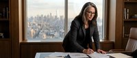 Professional businesswoman with glasses reviewing documents at executive office desk with city skyline view through window during daytime meeting.